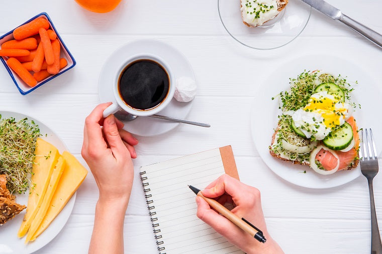A person grips a cup of coffee with one hand and is writing in a journal with the other, while surrounded by a variety of healthy foods. 