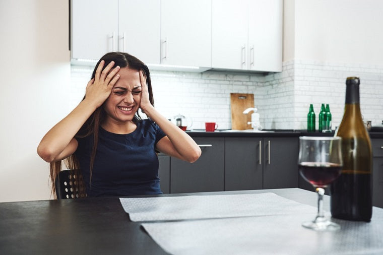 A young woman with long brown hair grips her head in pain. A wine bottle and glass of wine are on the table in front of her. 