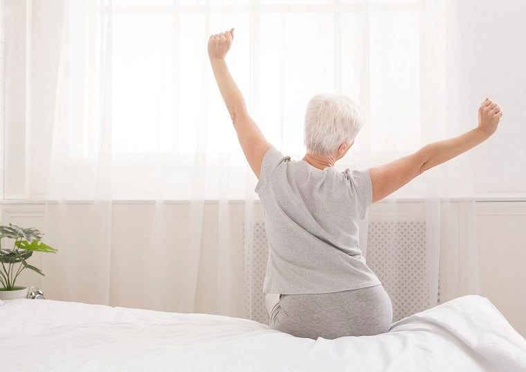 An older woman with short gray hair is on the edge of the bed, stretching her arms overhead in the morning sunlight filtering through the window after a great night of sleep. 