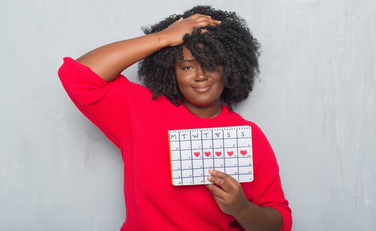 A woman wearing a red sweater and with brown hair shows what appears to be her menstrual cycle marked with little red hearts on a calendar 
