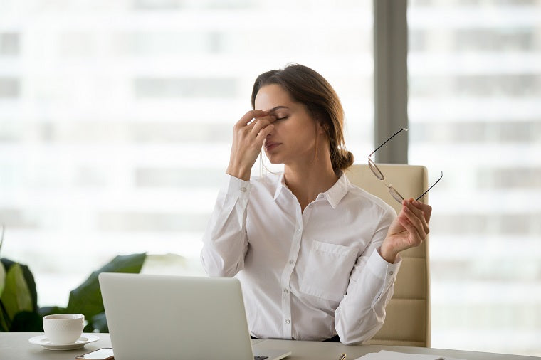 A professional woman in a white buttoned shirt has a laptop in front of her, has removed her glasses, closed her eyes and is pinching the bridge of her nose, clearly showing signs of pain from a migraine. 