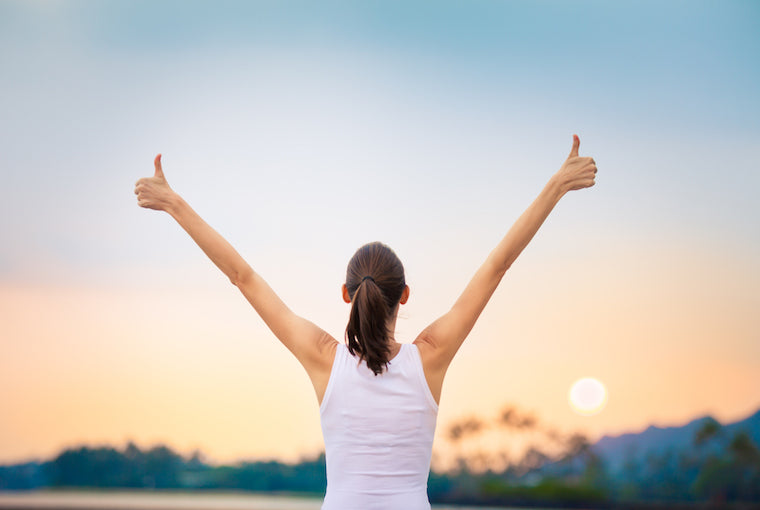 A woman observes a stunning, pastel sunrise with her back to the camera. Her arms are extended overhead and she is giving two thumbs up, suggesting she is joyful. 