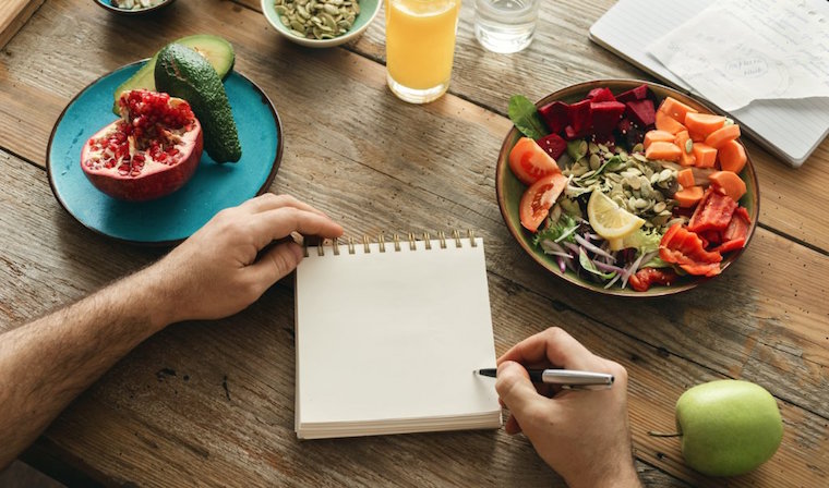 A person is writing on a notepad with fruit on a plate to their left, a salad to their right and a glass of juice in the middle. 