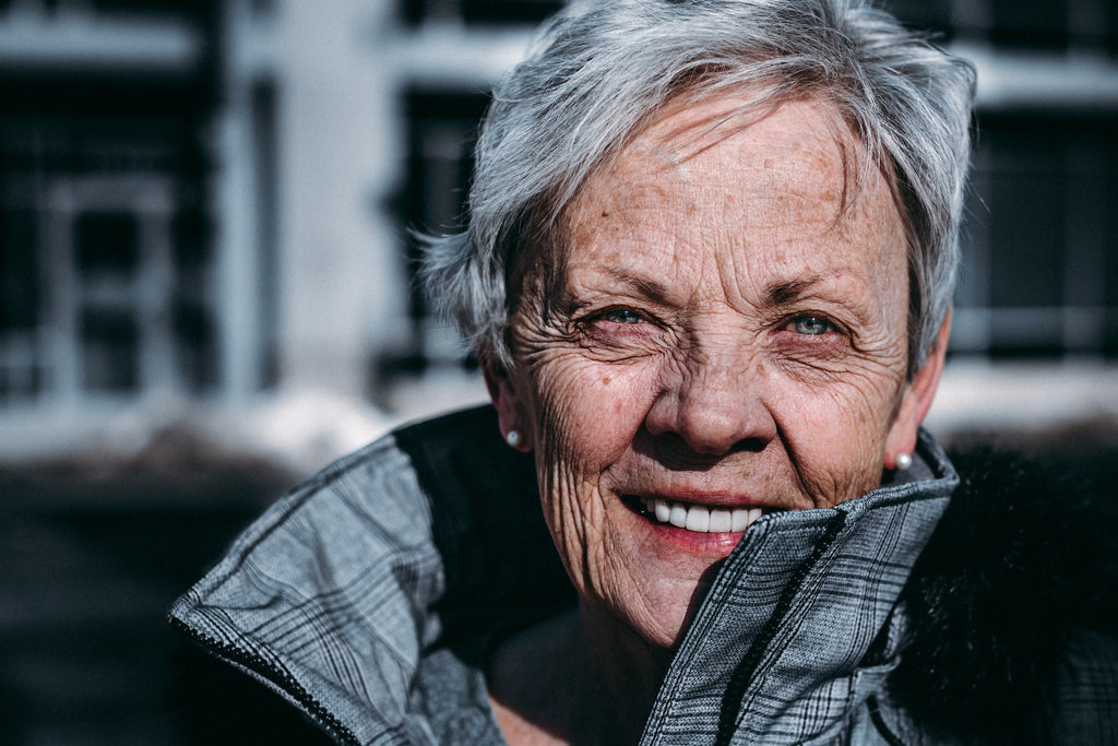 An older woman smiles happily and confidently into the camera, her jacket collar pulled up. 