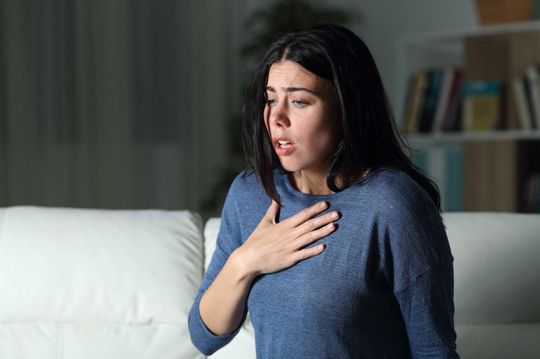 A woman in her 30s with long dark hair and wearing a blue shirt looks panicked and is holding her hand over her chest, like she is experiencing a panic attack. 